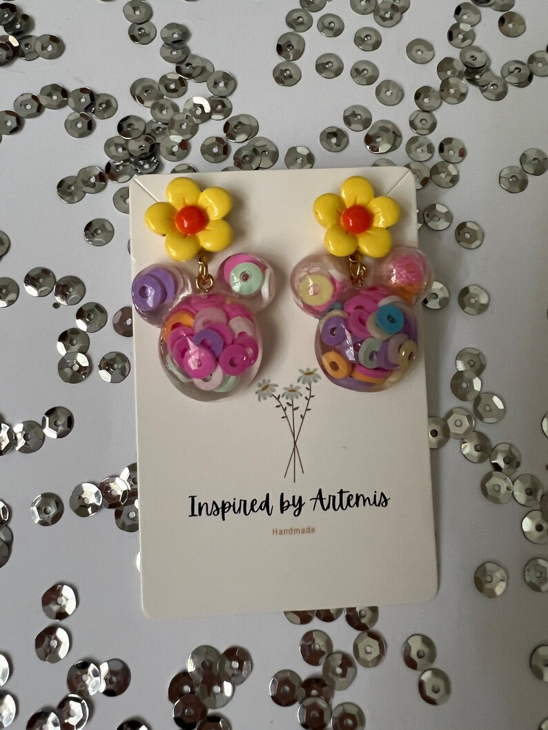 May include: A pair of handmade earrings featuring a yellow flower charm and a clear resin charm filled with colorful confetti. The earrings are labeled "Inspired by Artemis" and "Handmade".