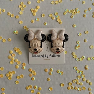 Puede incluir: Un par de pendientes hechos a mano con un personaje de dibujos animados con cabello negro, un lazo blanco y una cara amarilla. Los pendientes est&aacute;n en una tarjeta blanca con el texto "Inspired by Artemis" y "Handmade".