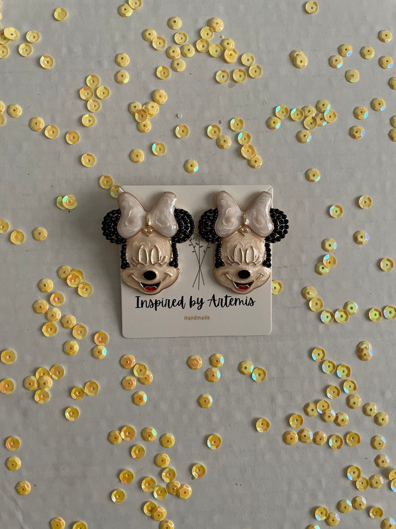 Puede incluir: Un par de pendientes hechos a mano con un personaje de dibujos animados con cabello negro, un lazo blanco y una cara amarilla. Los pendientes est&aacute;n en una tarjeta blanca con el texto "Inspired by Artemis" y "Handmade".