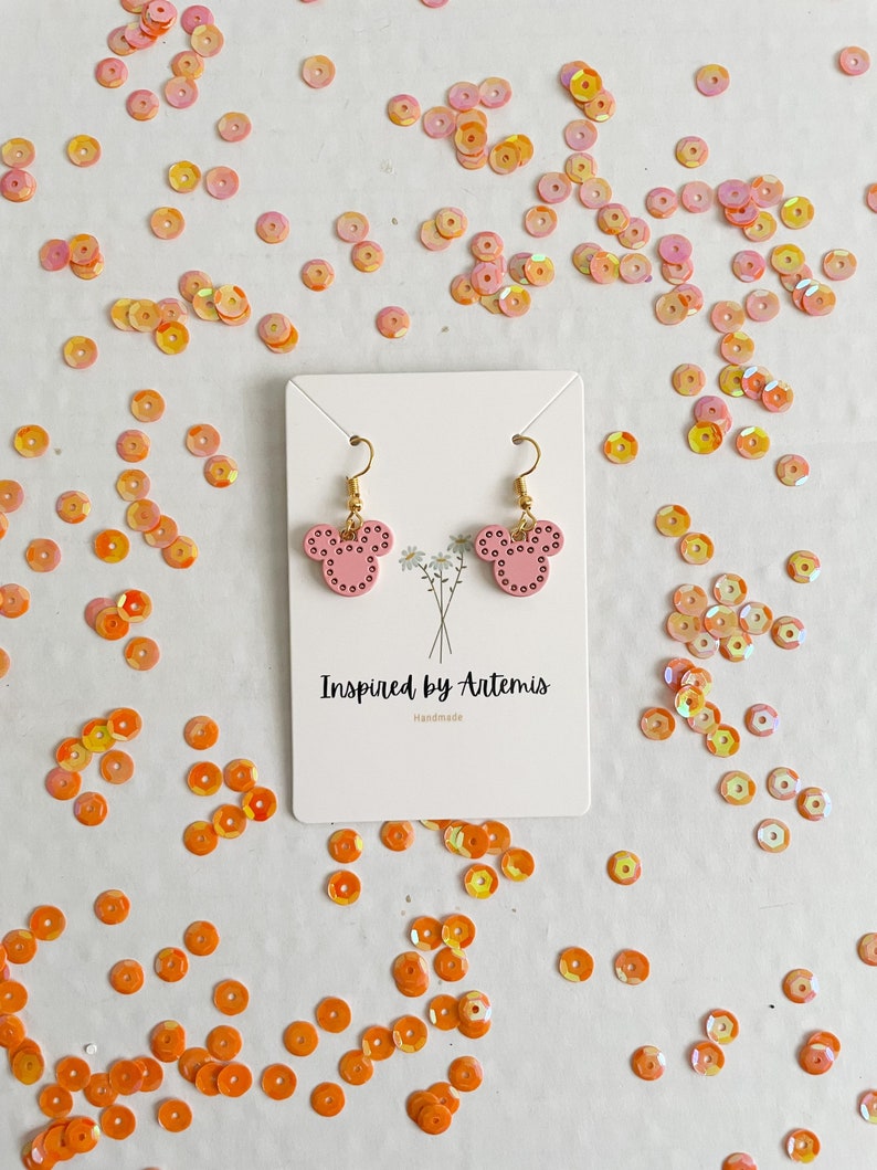May include: Pink mouse-shaped earrings with gold hooks and white polka dots. The earrings are on a white card with the text "Inspired by Artemis Handmade".
