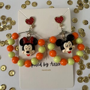 May include: A pair of dangle earrings featuring Mickey Mouse and Minnie Mouse characters. The earrings are made with orange and light green beads and have a red heart charm at the top.