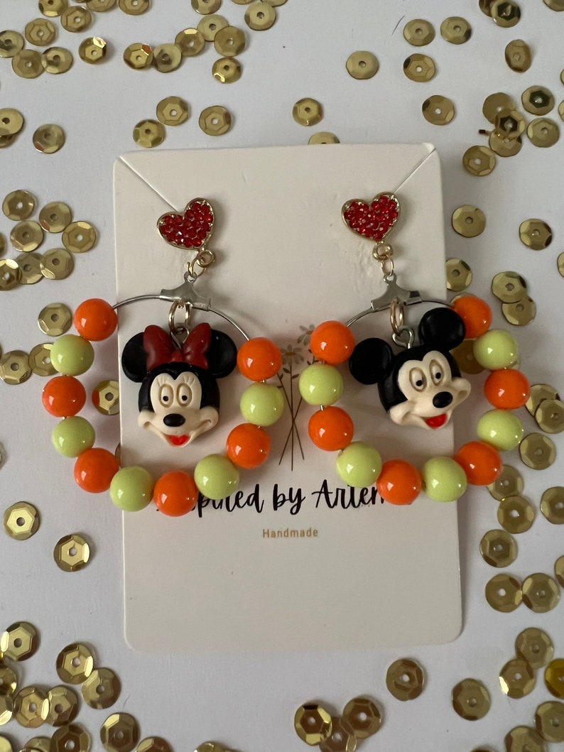 May include: A pair of dangle earrings featuring Mickey Mouse and Minnie Mouse characters. The earrings are made with orange and light green beads and have a red heart charm at the top.