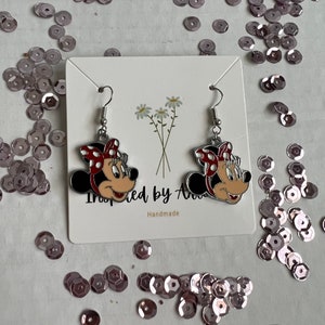 May include: A pair of silver earrings with red and white Minnie Mouse character charms. The earrings are displayed on a white card with the text "Inspired by Ava" and "Handmade".