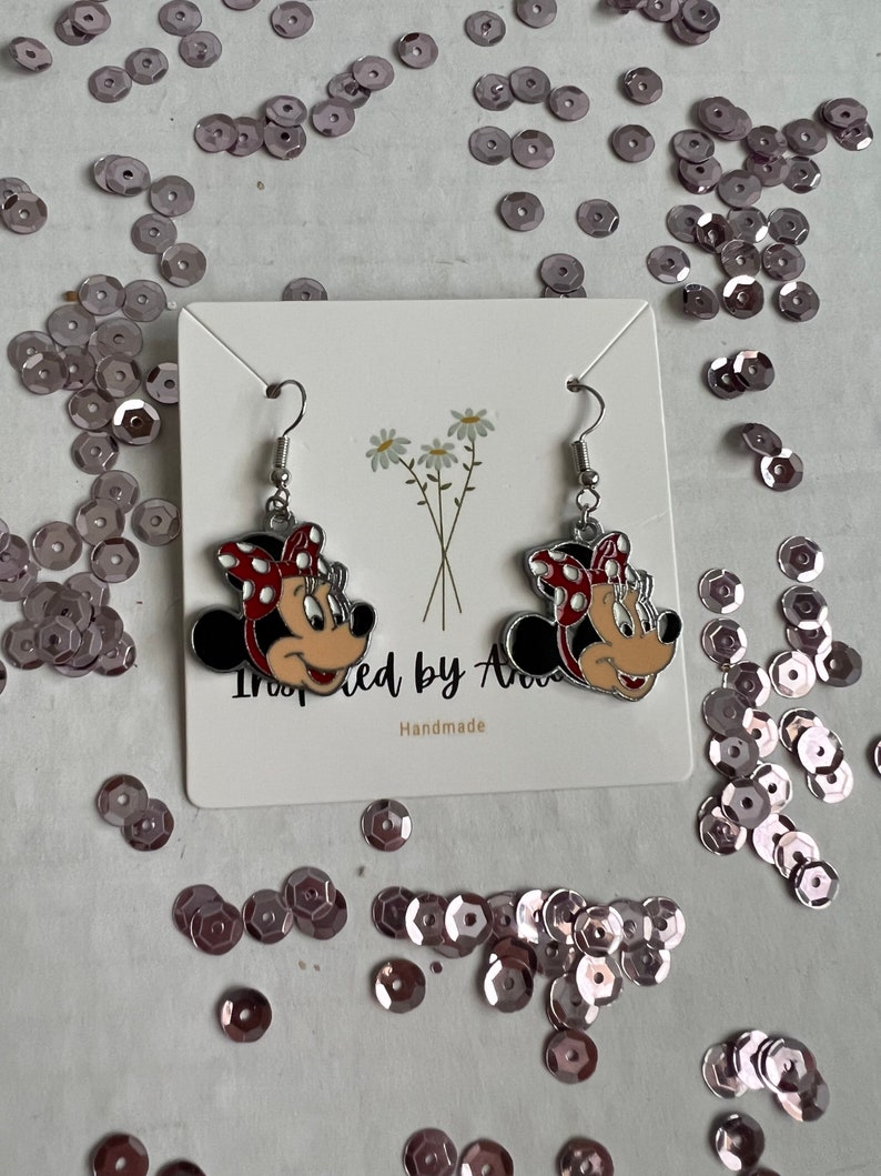 May include: A pair of silver earrings with red and white Minnie Mouse character charms. The earrings are displayed on a white card with the text "Inspired by Ava" and "Handmade".