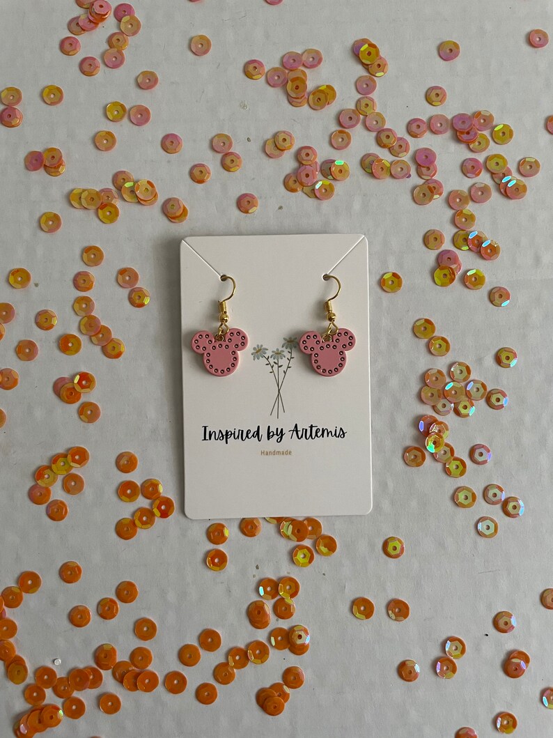 May include: Pink mouse-shaped earrings with gold hooks and white polka dots. The earrings are on a white card with the text "Inspired by Artemis Handmade".