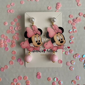 May include: Pink Minnie Mouse earrings with a pearl and pink ball charm. The earrings are handmade and feature a pink bow and a white background.