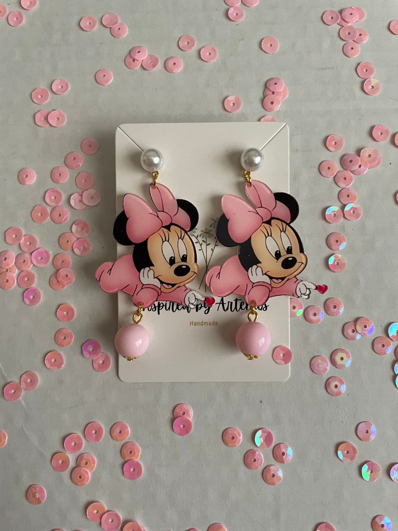 May include: Pink Minnie Mouse earrings with a pearl and pink ball charm. The earrings are handmade and feature a pink bow and a white background.