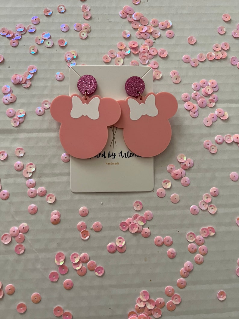 Puede incluir: Pendientes de color rosa en forma de Minnie Mouse con lazos blancos y detalles de purpurina. Los pendientes est&aacute;n sobre una tarjeta blanca con el texto "Wicked by Artemis" y "Handmade".