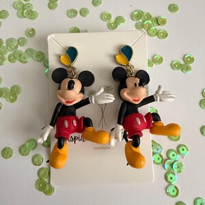 May include: A pair of Mickey Mouse earrings. Each earring features a black head with large ears, a red shorts, yellow shoes, white gloves, and a blue and yellow accent. The earrings are on a white card.