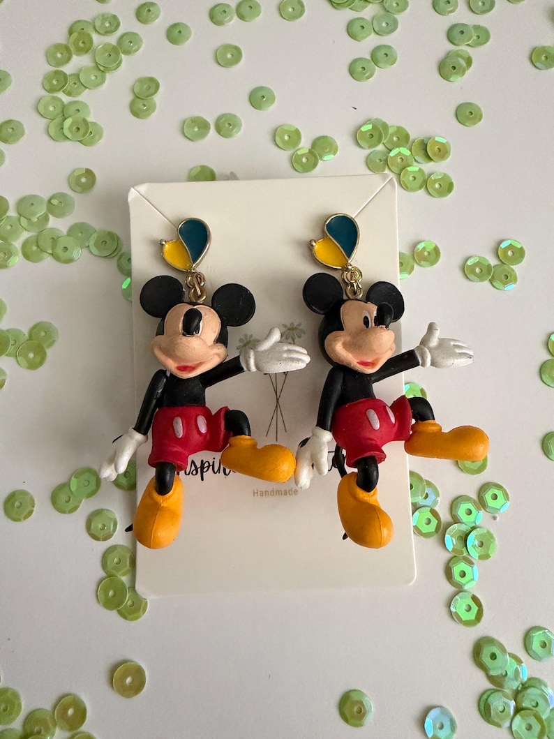 May include: A pair of Mickey Mouse earrings. Each earring features a black head with large ears, a red shorts, yellow shoes, white gloves, and a blue and yellow accent. The earrings are on a white card.
