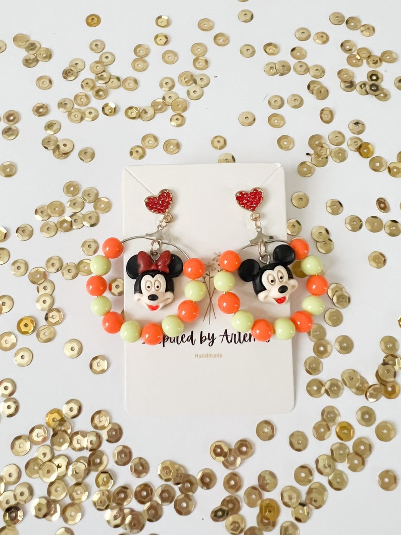 May include: A pair of dangle earrings featuring a red heart charm and a Mickey Mouse character charm. The earrings are made with orange and light green beads and are surrounded by gold sequins.