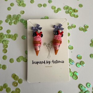 May include: A pair of handmade ice cream cone earrings with Minnie Mouse-inspired details. The earrings feature pink ice cream scoops, black mouse ears, red bows, and gray bows at the top. The earrings are on a white card with the text "Inspired by Artemis" and "Handmade".