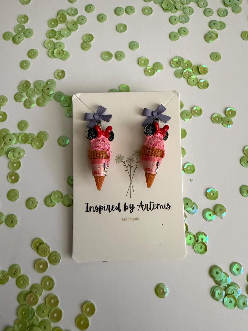 May include: A pair of handmade ice cream cone earrings with Minnie Mouse-inspired details. The earrings feature pink ice cream scoops, black mouse ears, red bows, and gray bows at the top. The earrings are on a white card with the text "Inspired by Artemis" and "Handmade".
