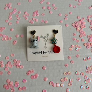 Peut inclure: Boucles d'oreilles faites &agrave; la main avec un personnage de dessin anim&eacute; et un motif floral. Une boucle d'oreille repr&eacute;sente un personnage gris et bleu, l'autre une fleur verte et un charme rouge. Les boucles d'oreilles sont sur une carte blanche avec le texte "Inspired by Arte".