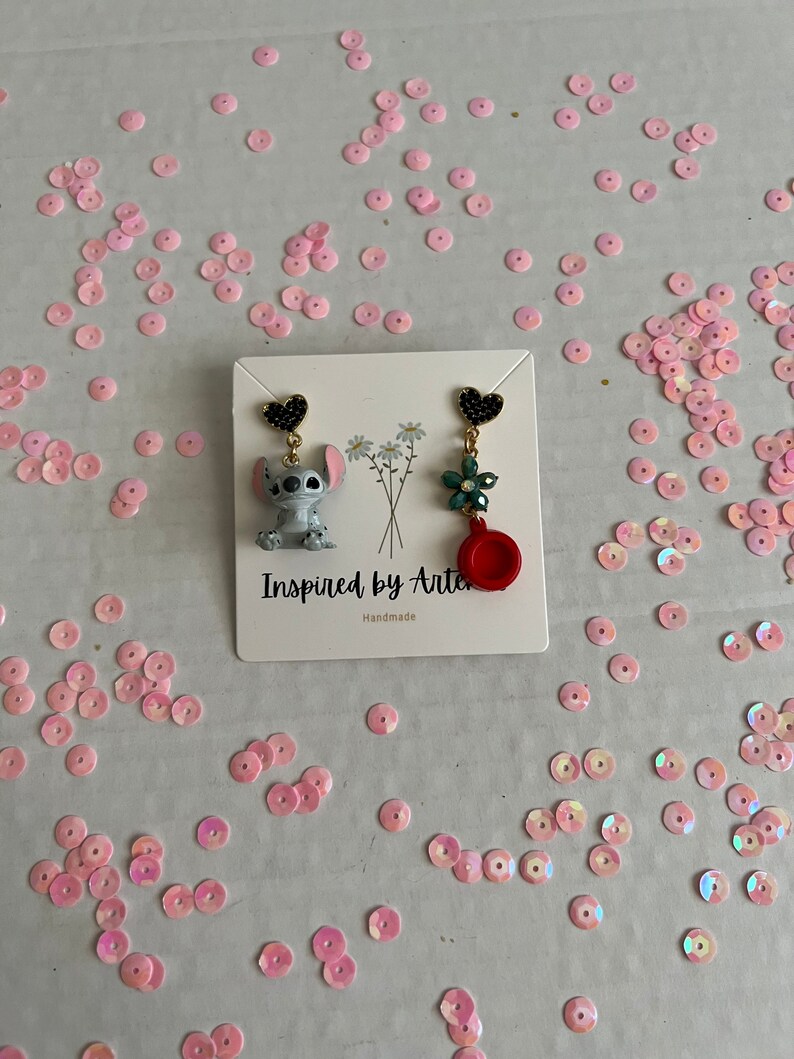 Peut inclure: Boucles d'oreilles faites &agrave; la main avec un personnage de dessin anim&eacute; et un motif floral. Une boucle d'oreille repr&eacute;sente un personnage gris et bleu, l'autre une fleur verte et un charme rouge. Les boucles d'oreilles sont sur une carte blanche avec le texte "Inspired by Arte".