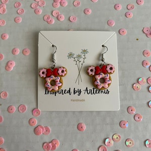 May include: A pair of donut-shaped earrings with pink frosting and sprinkles, red bows, and silver hooks. The earrings are displayed on a white card with the text "Inspired by Artemis" and "Handmade".