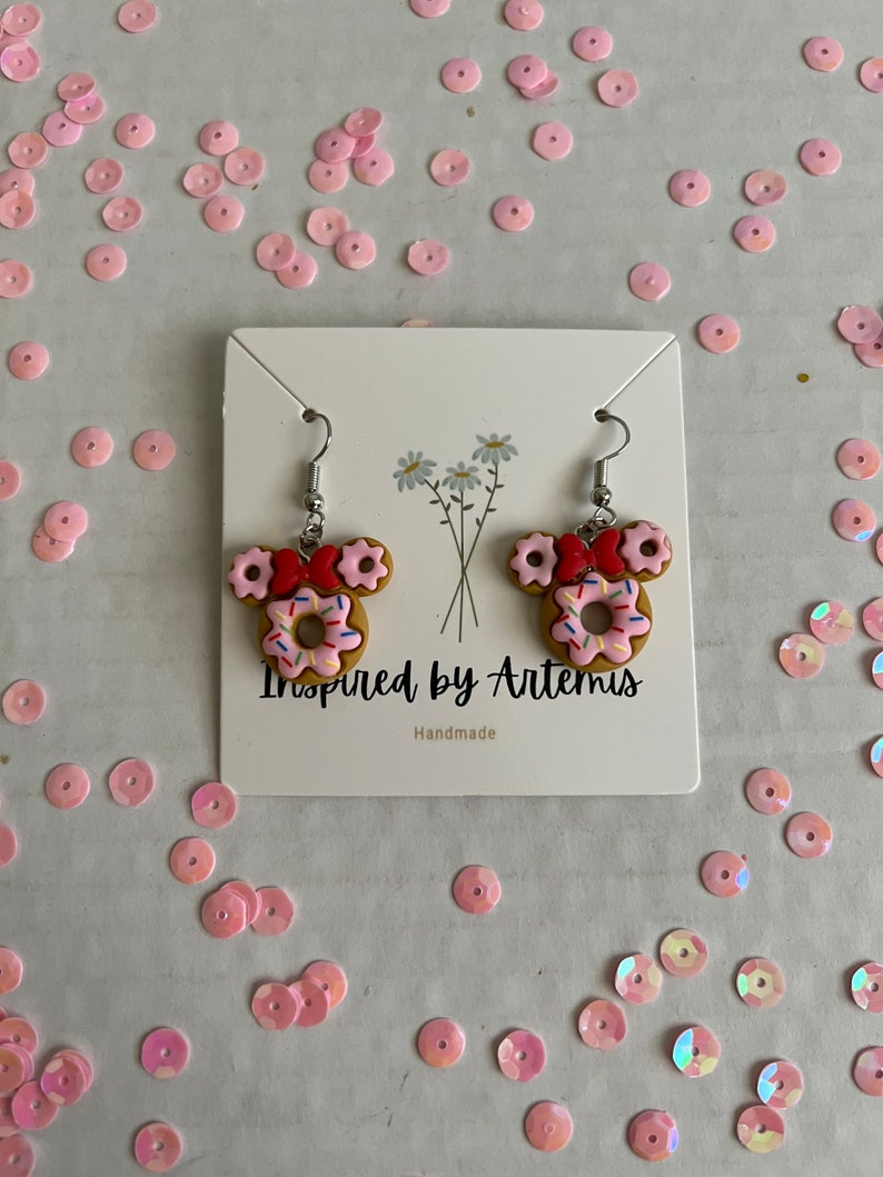 May include: A pair of donut-shaped earrings with pink frosting and sprinkles, red bows, and silver hooks. The earrings are displayed on a white card with the text "Inspired by Artemis" and "Handmade".