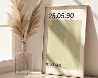 Könnte beinhalten: Ein gerahmter Druck mit dem Datum "25.05.90" und den Namen "Nina & Sam" in minimalistischem Design. Der Druck hat ein hellgrünes Rechteck auf weißem Hintergrund, in einem goldenen Rahmen. Eine klare Glasvase mit Pampasgras steht daneben.