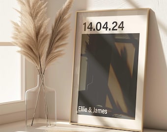 Könnte beinhalten: Ein gerahmter Druck mit dem Datum "14.04.24" und den Namen "Ellie & James" in weißer Schrift. Der Druck hat einen goldenen Rahmen und eine neutrale Farbpalette. Eine klare Glasvase mit Pampasgras befindet sich neben dem Druck.