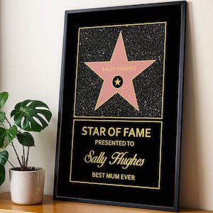 Puede incluir: Impresión enmarcada en negro con una estrella rosa con el nombre "SALLY HUGHES". El texto dorado debajo dice "STAR OF FAME" y "BEST MUM EVER". La impresión se muestra sobre una superficie de madera.