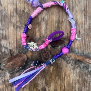 Op de afbeelding: Een handgemaakte haarwikkel met roze, paarse en blauwe draden. Versierd met bedels, kralen en linten, waaronder een maanbedel, een oogbedel en een kristal. Ontworpen om rond een vlecht of paardenstaart te wikkelen.