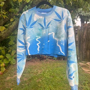 May include: A light blue cropped sweatshirt featuring a white spray-painted effect and a repeating pattern of dark blue cannabis leaf silhouettes. The garment has long sleeves, a crew neckline, and a small Champion logo on the left sleeve cuff.