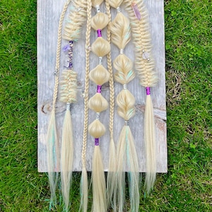 Puede incluir: Extensiones de cabello rubio con elementos decorativos. Las extensiones están dispuestas sobre una tabla de madera sobre un fondo de hierba verde. Las extensiones presentan trenzas, cuentas y detalles morados. Algunos mechones tienen vetas azules y verdes.