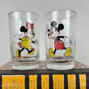 May include: Two clear glass drinking glasses with cartoon images of Mickey Mouse and Minnie Mouse. Mickey Mouse is wearing red shorts and yellow shoes. Minnie Mouse is wearing a yellow dress and red shoes. The glasses are sitting on top of a stack of books.