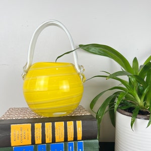 May include: A yellow glass vase with a white handle and a swirl design. The vase is sitting on top of a stack of books. A green plant in a white pot is to the right.