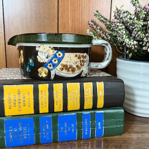May include: A ceramic gravy boat with a dark brown exterior, a green rim, and a white handle. The boat features a whimsical design with a figure wearing a blue collar with yellow flowers. It rests on a stack of vintage books.