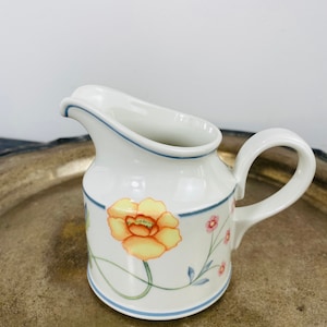 May include: White ceramic creamer with a floral design. The creamer features a yellow and orange flower with pink and blue accents. A blue line borders the top and bottom of the creamer.