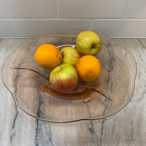 Può includere: Una fruttiera di vetro trasparente piena di frutta fresca. La fruttiera contiene due arance e tre mele. Le mele sono verdi e rosse, e le arance sono arancioni brillanti. La fruttiera è su un piano di lavoro in marmo.