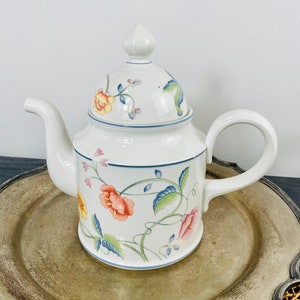 May include: White ceramic teapot with a lid, featuring a floral design in shades of orange, pink, yellow, and blue. The teapot has a curved spout and a large handle. The design includes a blue trim.
