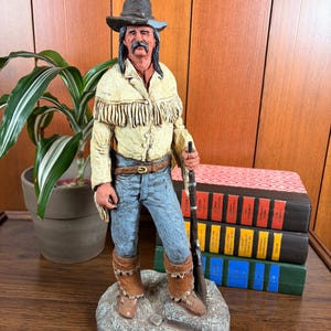 May include: A detailed figurine of a frontiersman, wearing a hat, fringed jacket, and blue jeans, holding a rifle. The figure stands on a textured base. The color palette includes tan, brown, and blue tones. The figurine is a decorative item.