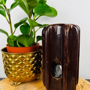 May include: A dark brown ceramic vase with vertical ridges and a circular opening. The vase is next to a small potted plant in a gold-colored, textured planter. The plant has bright green leaves.