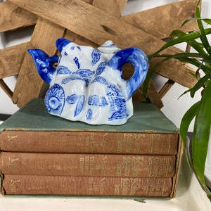 Op de afbeelding: Kleine, wit en blauwe porseleinen theepot met een gedetailleerd ontwerp. De theepot is versierd met blauwe patronen en figuren. Hij staat bovenop een stapel vintage boeken met bruine kaften en groene ruggen. Een houten stervormige decoratie op de achtergrond.