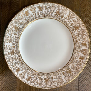 Puede incluir: Un plato de cena de cerámica blanca con un borde dorado y un intrincado diseño floral y de pájaros dorado.