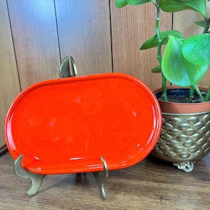 Puede incluir: Un plato de cerámica naranja brillante, de forma ovalada, expuesto sobre un soporte de metal dorado. El plato tiene un acabado brillante. Una planta en maceta con hojas verdes es visible en el fondo.