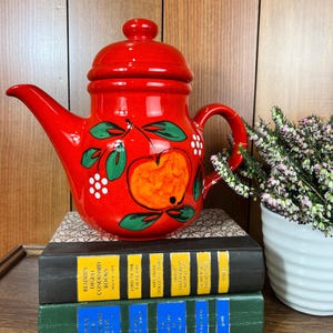 May include: A vibrant red ceramic teapot with a lid, adorned with a hand-painted apple design. The teapot sits atop a stack of vintage books with gold and yellow lettering. A small white pot with purple flowers is to the right.