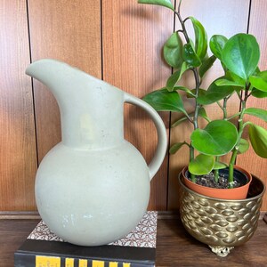 Puede incluir: Una jarra de cerámica gris claro con un cuerpo redondeado y un pico curvo. La jarra está sentada sobre una pila de libros con una cubierta de patrón marrón y blanco. Una planta en maceta con hojas verdes está en el fondo.