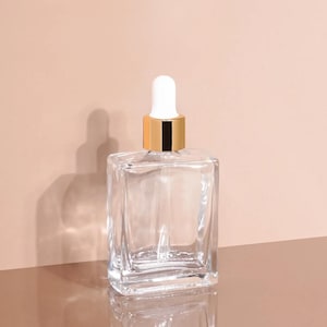May include: A clear, square glass bottle with a white dropper and gold-coloured collar. The bottle is empty and sits on a reflective surface, casting a shadow against a neutral-toned background. The design is minimalist and elegant.