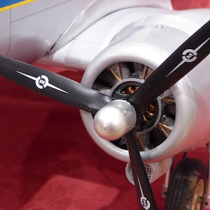 May include: Close-up of a silver aircraft engine with a black propeller. The propeller has a white "N" logo on each blade.