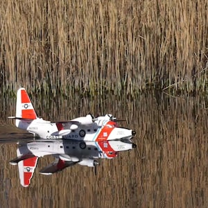 May include: A white and red Coast Guard aircraft with the text "COAST GUARD" on the side is floating on a body of water. The aircraft is reflected in the water.