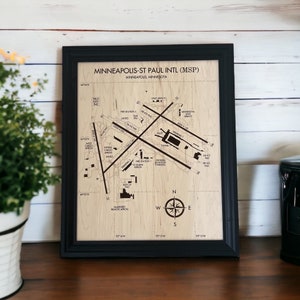 May include: Framed wooden map of Minneapolis-St Paul International Airport (MSP). The map features runways, terminals, and other airport infrastructure, with text labels. The frame is black, and the map is set against a light wood grain background. A compass rose is visible.