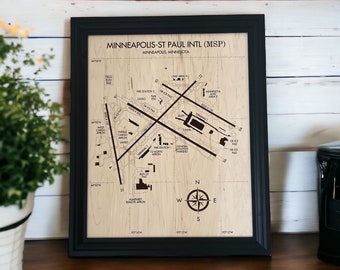 Può includere: Mappa in legno incorniciata dell'aeroporto internazionale di Minneapolis-St Paul (MSP). La mappa presenta piste, terminal e altre infrastrutture aeroportuali, con etichette di testo. La cornice è nera e la mappa è su uno sfondo di venature del legno chiaro. È visibile una rosa dei venti.