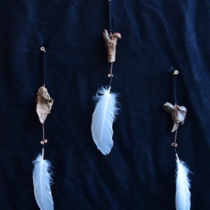 Puede incluir: Tres adornos colgantes con plumas blancas y objetos óseos marrones envueltos en alambre de cobre.