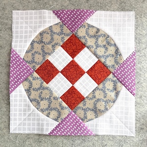 May include: Square quilt block featuring a central red and white diamond pattern, surrounded by a circular design with floral print fabric in beige and blue. Accented with purple polka dot triangles and a white grid background.