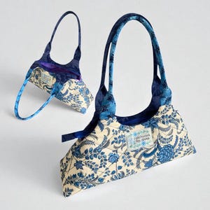 May include: Two small, quilted handbags with blue floral patterns on an off-white background. The bags have dark blue handles and accents. One bag is in the foreground, and the other is in the background. The bags have a label that says "patchwork by anna".