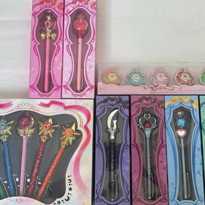 May include: A collection of Sailor Moon themed pens and accessories in their original packaging. The set includes two vertical pink boxes with wands, a horizontal box with five colorful pens, and four individual vertical boxes containing wands.