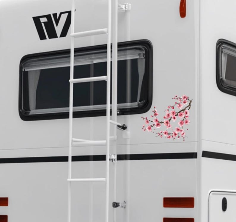 May include: White RV exterior with a black window, ladder, and cherry blossom decal. The decal features pink flowers and brown branches. The RV has black stripes and a red light.